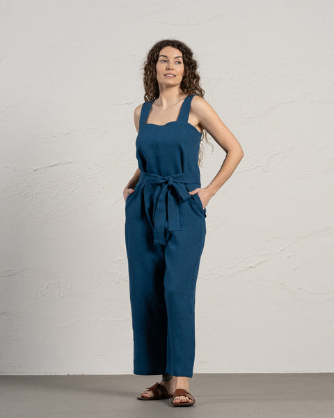 A person with light skin and long, dark curly hair is standing with their hands in the pockets of a linen jumpsuit. The jumpsuit has wide straps and a tied belt at the waist, and they are wearing brown sandals.