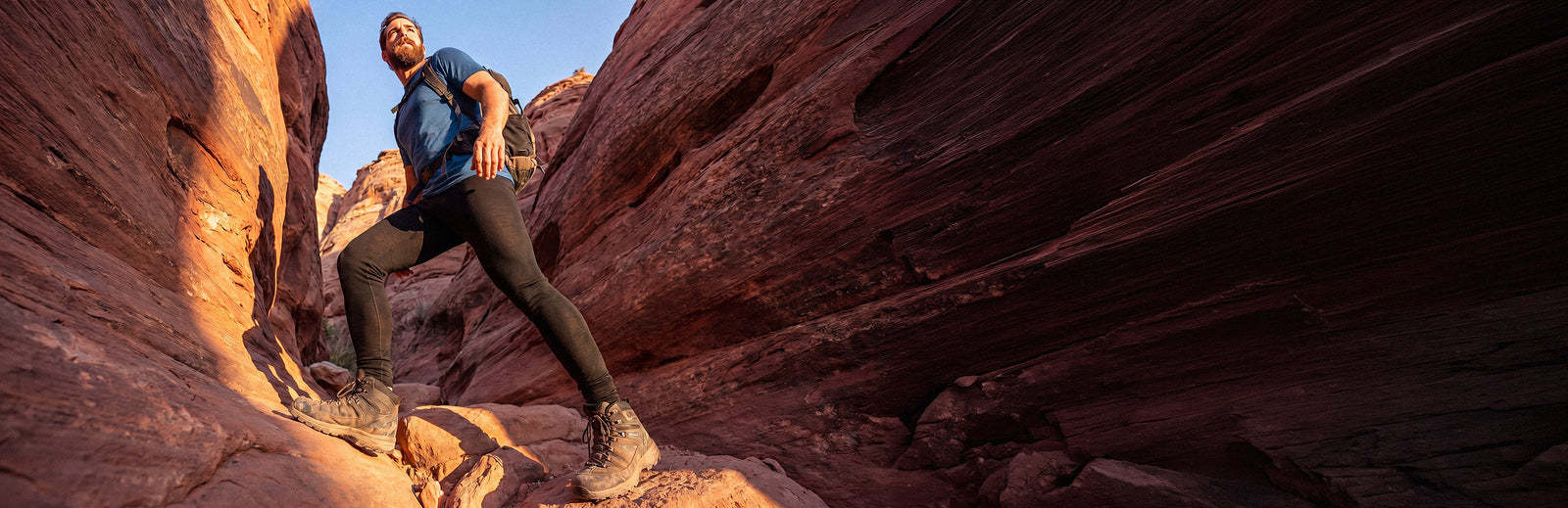 A low-angle outdoor lifestyle shot of a bearded man hiking up a rugged red rock canyon. He is captured mid-stride, stepping upward on the uneven terrain. He is wearing a short-sleeved blue t-shirt, black athletic leggings, sturdy brown hiking boots, and carrying a backpack. The warm, golden hour sunlight casts shadows and highlights the deeply striated texture of the canyon's rock walls. A narrow strip of clear blue sky is visible through the opening at the top left.