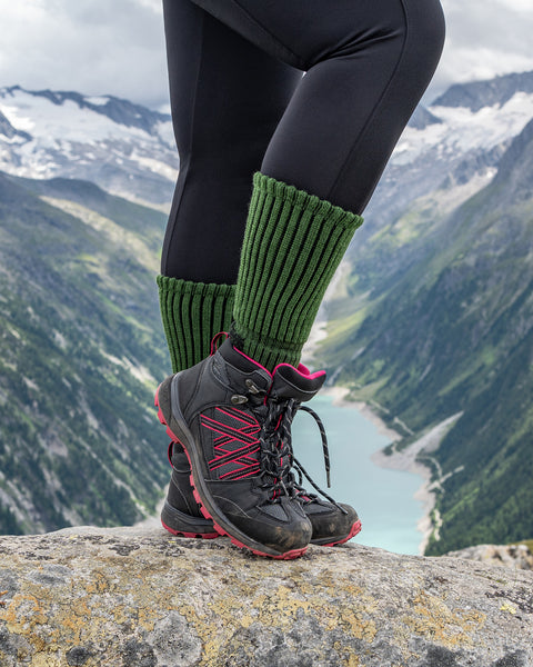 Close-up of a hiker wearing green ribbed Merino hiking socks with black and pink hiking boots, standing on a rock overlooking an alpine lake and mountains.