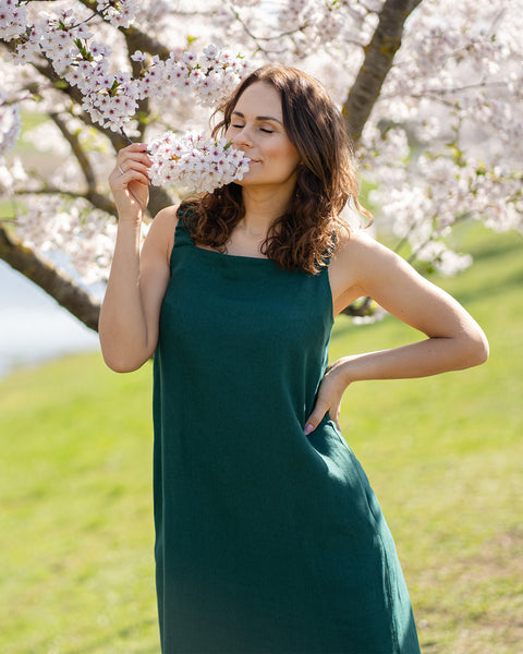A woman with brown hair, wearing a long, dark green, sleeveless maxi dress, stands outdoors next to a tree with white blossoms.