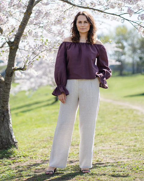 A woman with fair skin and dark, wavy hair, wearing a shadow puprle, off-the-shoulder top, is shown from the waist up. She is looking directly at the viewer with a slight smile.