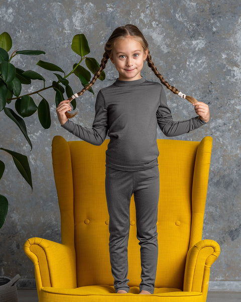 Smiling girl in gray Merino wool base layer set standing on a yellow armchair, playing with her braids.