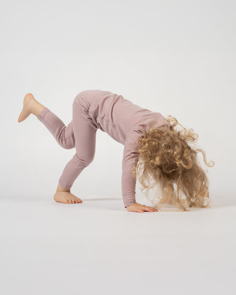 Child in merino wool dusty pink outfit performing a backbend on a white background