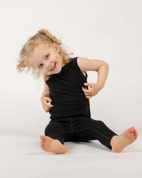 A young child with light curly hair in a ponytail is sitting on a white surface against a plain white background. They are wearing a black sleeveless top and black leggings. The child is looking at the camera with a wide, joyful smile, and their head is tilted slightly. Their bare feet are visible, and their hands are touching their top.