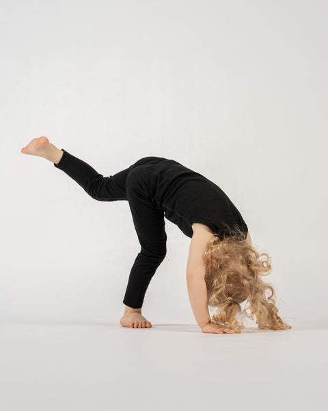 A young child with long, curly blonde hair is captured in a dynamic pose against a plain white background. They are wearing a black short-sleeved top and black leggings. The child is bent over, with their hands on the ground and one leg extended straight up behind them, resembling a handstand or a yoga pose. Their face is not visible.