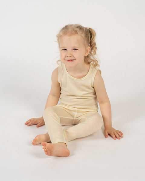 Young child wearing a merino sleeveless top and pants in the color natural sitting on a white background