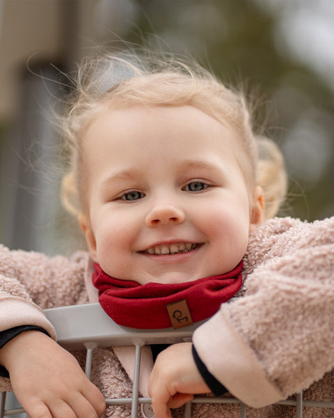 A smiling child with blonde pigtails and blue eyes looks happily at the camera, wearing a red Menique neck gaiter and a cozy beige jacket, leaning over a metal railing. The child's joyful expression fills the frame.