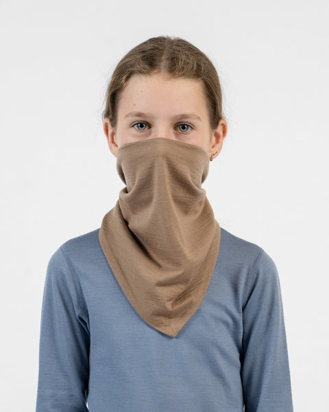 A young girl wearing a beige merino bandana pulled up to cover her nose, mouth, and neck, paired with a blue long-sleeve shirt.