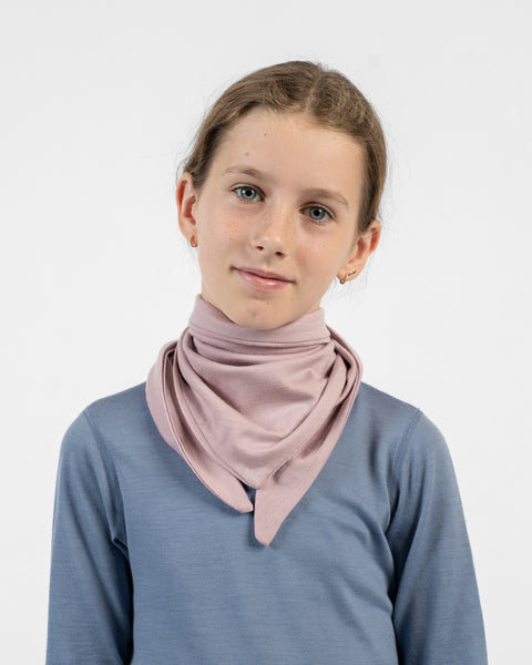 A young girl wearing a dusty pink merino bandana draped loosely around her neck like a cowl, over a blue long-sleeve shirt.
