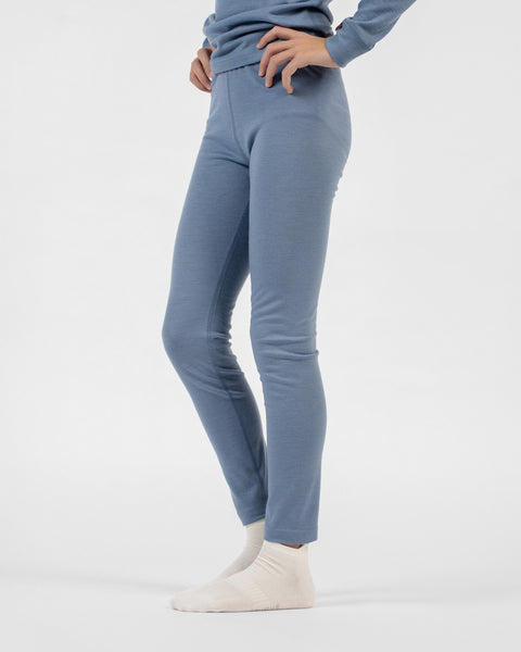 A close-up side view of a child's lower half wearing fitted dusty blue merino bottoms and cream-colored socks, with hands resting near the waistband.