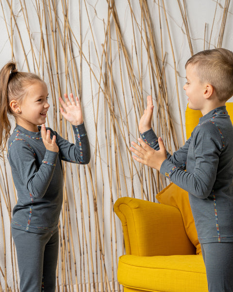 Smiling boy and girl wearing matching grey Merino wool base layer sets with colorful seams, playing a clapping game next to a bright yellow armchair.