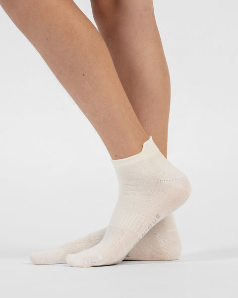 A side-profile view of a person's lower legs and feet wearing natural white cotton ankle socks, with the back foot slightly lifted on its toes to reveal branding on the sole.