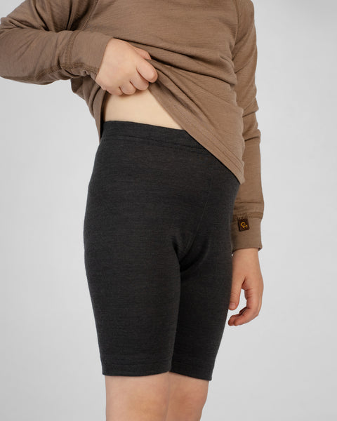 A slightly angled view of a child wearing dark charcoal merino wool cycling shorts paired with a brown long-sleeve top. The child is lifting the front of the shirt with one hand to display the high-waisted fit of the shorts, set against a plain light gray studio background.