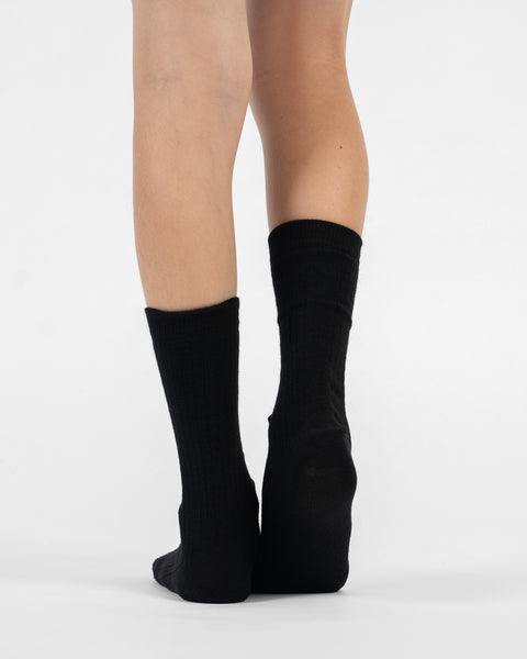 A back view of a person's lower legs and heels, standing flat in black ribbed merino crew socks.