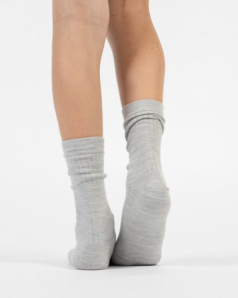 A back view of a person's lower legs and heels, standing flat in light gray ribbed merino crew socks.