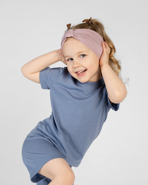 A studio portrait of a young girl leaning to her left with a playful smile. She is wearing a short-sleeved top and shorts set in blue, with a dusty pink ribbon headband. She has both hands near her head against a plain light grey background.