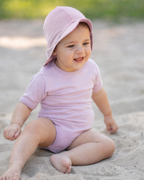 A smiling baby sitting barefoot in the sand outdoors, looking happily off to the side. The baby is wearing a light pink short-sleeve bodysuit and a matching dusty pink sun cap with a front brim and protective neck flap.