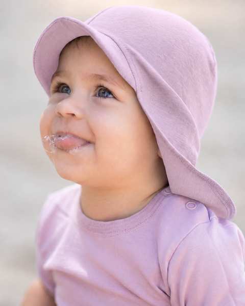 A close-up of a happy baby looking upwards with a little drool around their mouth, wearing a dusty pink short-sleeve top with shoulder snaps and a matching dusty pink sun cap featuring a front brim and neck cover.