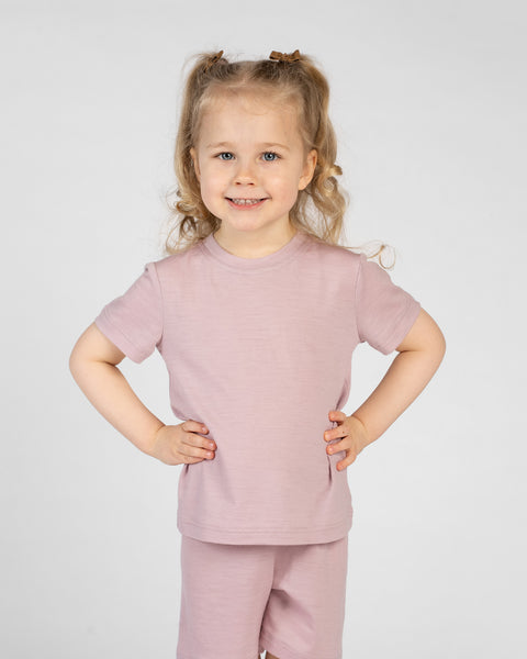 A young, smiling child stands against a light gray background wearing a dusty pink short-sleeve t-shirt and matching shorts. Their hands are placed on their hips, and their blonde hair is styled in two small pigtails with brown clips.