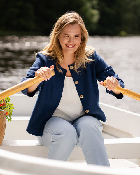 A woman wearing a blue jacket actively rows a small boat across a calm river. She holds the wooden oars firmly while looking forward and smiling.