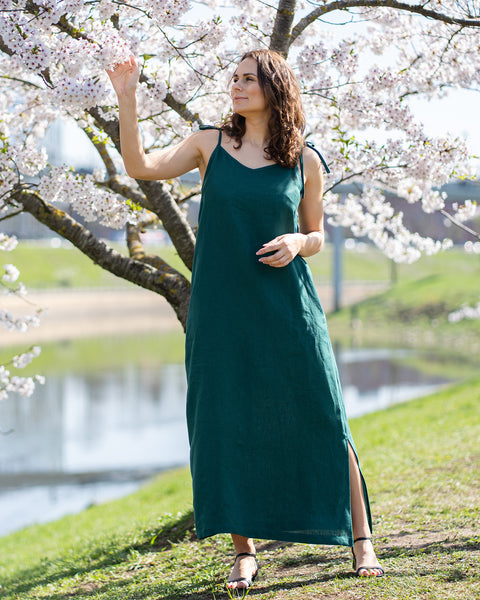 A woman with brown hair, wearing a long, dark green sleeveless dress with tie straps, stands outdoors next to a tree with white blossoms, touching the flowers.