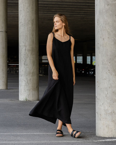 A light-skinned person with long blonde hair is standing in what appears to be an urban or industrial setting with large concrete pillars. They are wearing a flowing linen black sleeveless dress with thin straps and dark-colored sandals, and are looking off to the left.