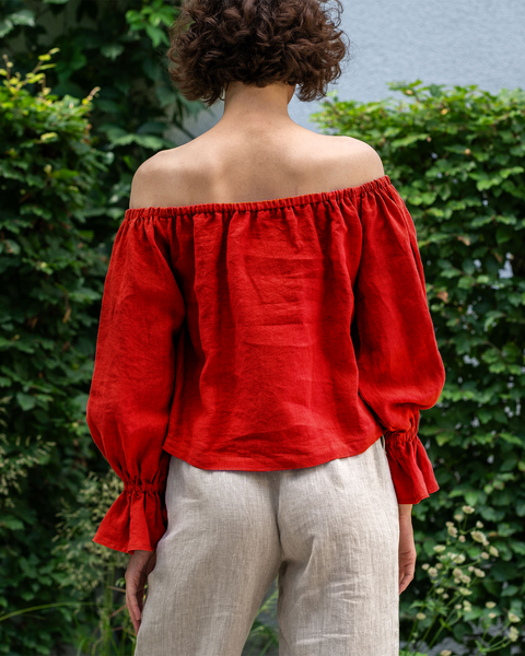 A woman with short, curly brown hair standing in front of a green ivy-covered wall. She is wearing a pure red off-the-shoulder linen top with voluminous sleeves that gather at the wrists, and light-colored pants.