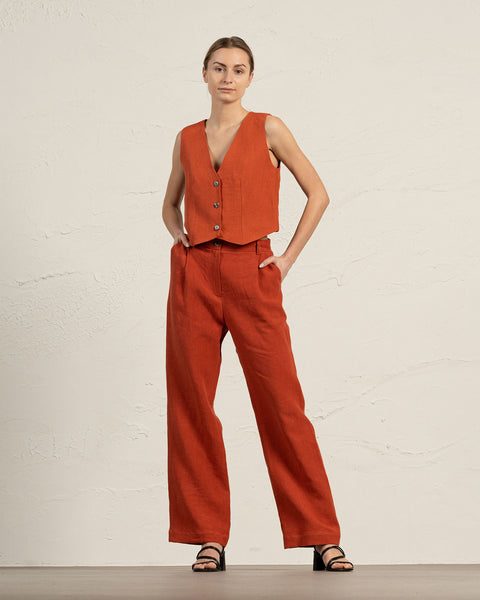 A fair-skinned woman models a cinnamon red colored linen vest and wide-leg trousers, standing against a textured light beige wall. She is wearing black sandals.