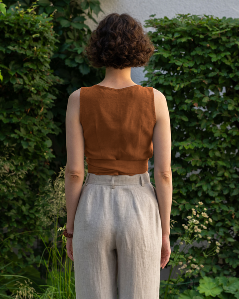 From the back, a light-skinned person with short, curly brown hair is standing outdoors. They are wearing an almond brown sleeveless wrap top and menique high-waisted pants. Lush green foliage is visible in the background.