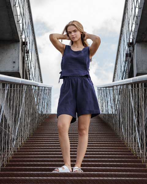 A full-body shot of a young woman with long, blonde hair standing at the bottom of a metal staircase, looking directly at the camera. She is wearing a storm blue linen two-piece outfit consisting of a sleeveless top with a scoop neck and side ties, and matching shorts with an elastic waistband. Her hands are placed behind her head. She is wearing white slide sandals. The staircase has metal steps and railings, and a concrete structure is visible above. The sky is cloudy.