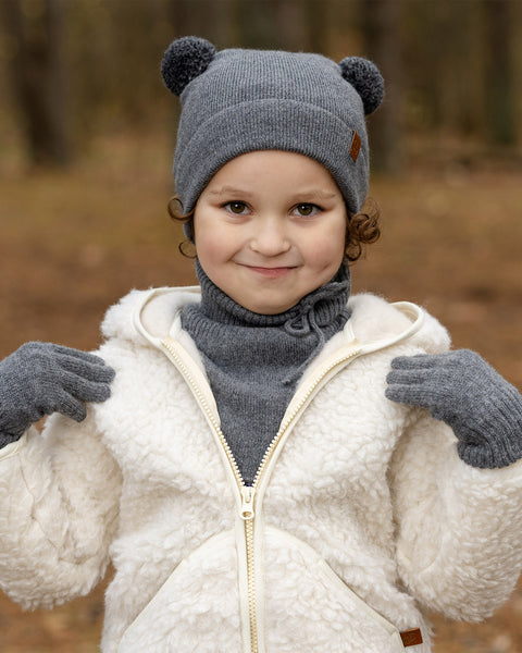 A young child with light brown curly hair and a subtle smile, is looking directly at the viewer. They are wearing a dark grey knit hat with two pom-pom ears and a brown tag, matching dark grey mittens, a dark grey turtleneck or neck warmer, and a cream-colored sherpa jacket with a zipper. The background is a blurred forest.