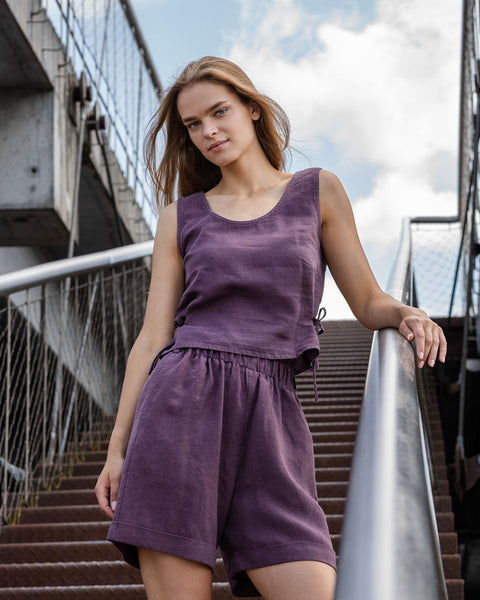 A full-body shot of a young woman with long, light brown hair standing on a metal staircase outdoors. She is looking directly at the camera with a neutral expression. She is wearing a two-piece linen outfit in a shadow purple color, consisting of a sleeveless top with a scoop neck and high-waisted shorts with an elastic waistband. Her left arm is resting on the metal handrail beside her. The staircase has textured, possibly rusted, steps.