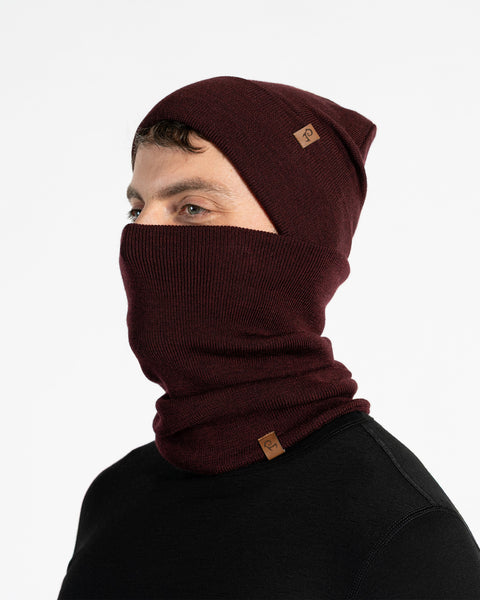 A male model, viewed in profile, wears a royal cherry beanie and a matching neck gaiter that covers his nose and mouth. Both the beanie and gaiter feature a small, brown leather patch with a Menique logo. He has a short beard and dark hair. The background is solid white.