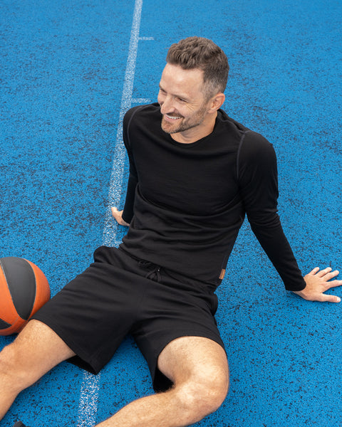 A smiling man sitting casually on a bright blue outdoor sports court, leaning back on one hand. He is wearing a black long-sleeved athletic top and black shorts, with an orange and black basketball resting on the ground next to his leg.