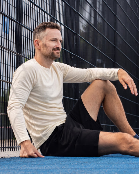 A man wearing a long-sleeved cream shirt and black shorts, sitting casually on a bright blue sports court next to a dark wire fence.