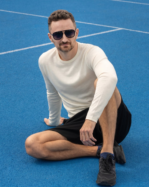 A man wearing dark sunglasses, a long-sleeved cream shirt, and black shorts, sitting on a bright blue sports court looking directly at the camera.
