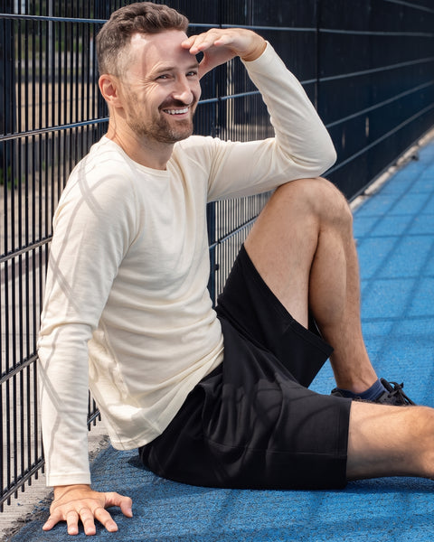 A man in a cream long-sleeved shirt and black shorts sitting on a blue athletic court, resting against a wire fence and using his hand to shield his eyes from the sun.