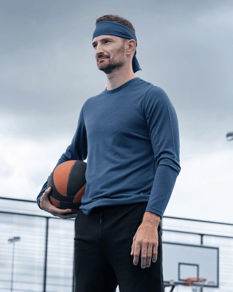 A man standing on an outdoor basketball court holding a basketball under his arm, wearing a dark blue long-sleeved shirt, black shorts, and a blue headband.