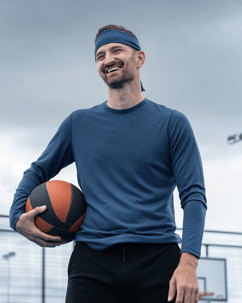 A slightly closer shot of man with a blue headband and dark blue long-sleeved shirt smiling on an outdoor basketball court while holding a basketball.