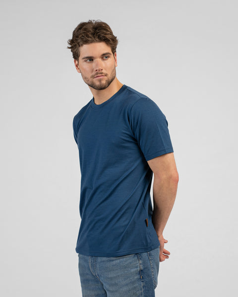 A man stands facing slightly away from the camera, looking back over his right shoulder with his hands clasped behind his back. He wears a denim blue short-sleeved t-shirt and blue jeans.