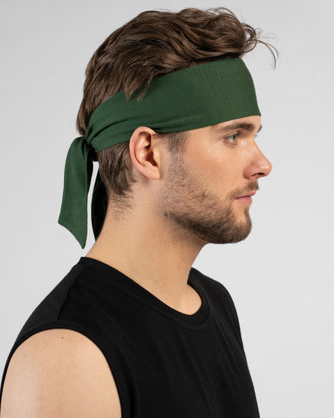 A side profile portrait of a man wearing a dark green tie headband and a black tank top. He faces the right against a plain light grey background.