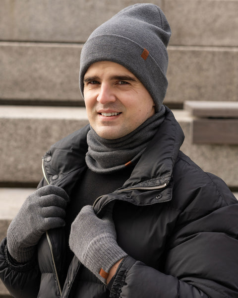 A smiling man with short dark hair and stubble wears a dark gray beanie, a matching gray neck gaiter, a black puffer jacket, and gray gloves. He is outdoors with concrete stairs in the background.