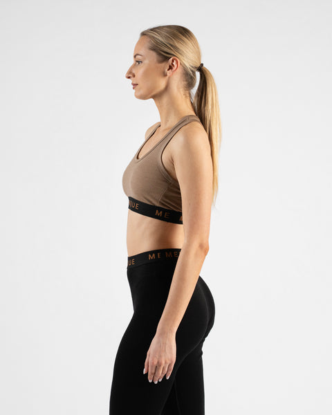 A side-profile studio shot of a blonde woman with her hair tied back in a ponytail, facing left. She is wearing a beige sports bra and black leggings, both featuring a black elastic band with the "MENIQUE" logo.