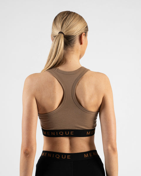 A back view studio shot of a blonde woman with a ponytail, showcasing the racerback design of a beige sports bra. She is also wearing black leggings, with both garments featuring a black elastic band with the "MENIQUE" logo against a plain white background.