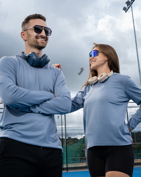 A man and a woman standing together on an outdoor sports court wearing matching light blue long-sleeved shirts, black shorts, and sunglasses.