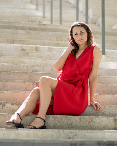 A woman with brown hair, wearing a bright red, sleeveless wrap dress, poses on indoor concrete stairs with metal railings.