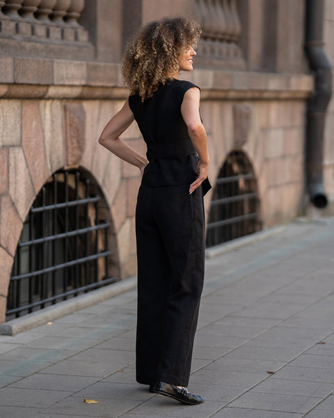 A person with voluminous curly hair, wearing a menique pure black sleeveless wrap top and wide-legged pure black trousers, stands in profile looking to the left with one hand in their pocket and the other on their hip. They are on a paved sidewalk next to a textured stone building with arched windows and a downspout.
