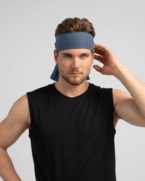 A fair-skinned man with brown hair and a short beard is wearing a black sleeveless shirt and a denim-blue tie headband. He is looking directly at the camera with his left hand raised to his hair, and the tied ends of the headband are visible behind his neck against a light gray background.