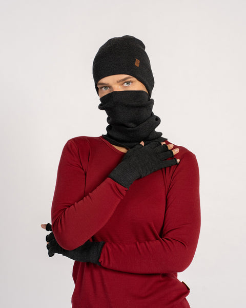 Frontal shot of a woman in a dark-gray beanie and gaiter, one hand touching her neck, wearing matching fingerless gloves.