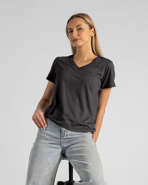 A studio portrait of a blonde woman sitting on a stool and looking directly at the camera. She is leaning back slightly, with her left hand resting in her lap and her right hand touching her hip. She is wearing a regular fit, charcoal grey short-sleeved v-neck t-shirt and light-wash blue jeans. The background is a solid light grey.